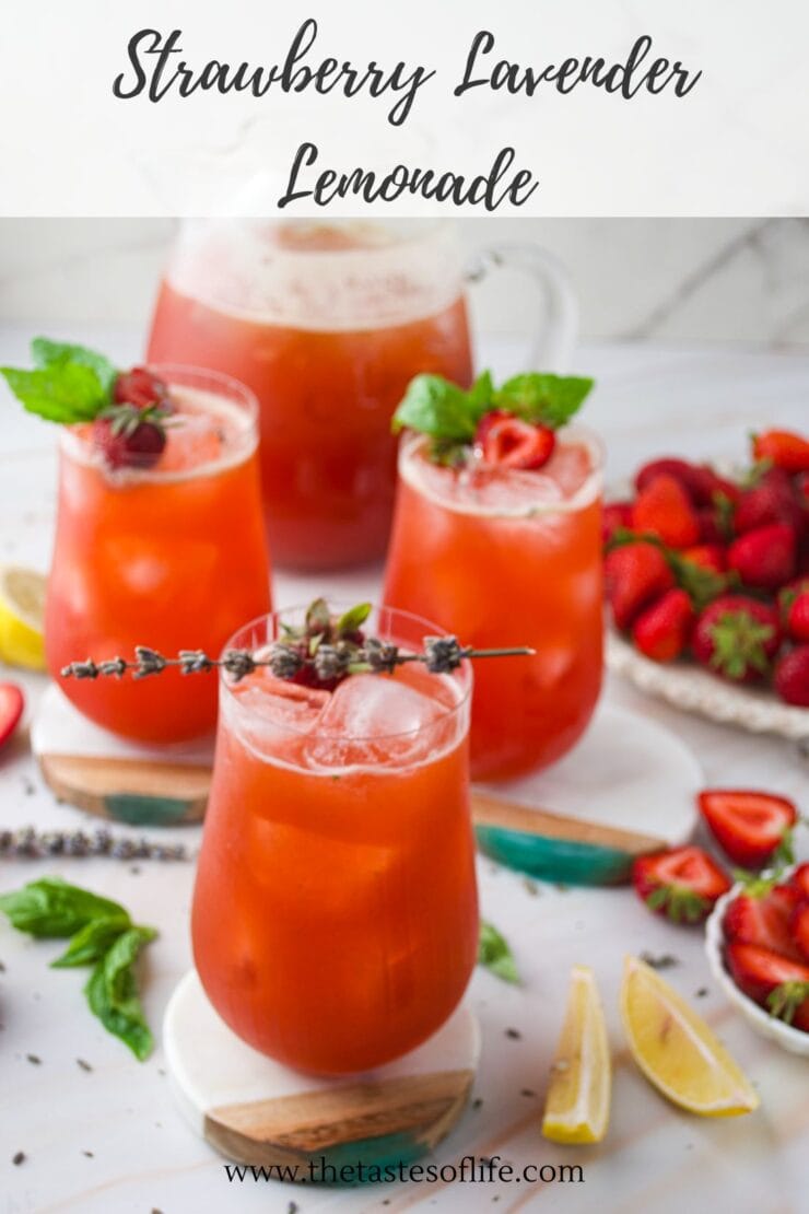 Four glasses of strawberry lavender lemonade with ice, garnished with strawberries, lavender sprigs, and mint. Fresh strawberries, lemon wedges, and mint leaves are scattered on a white surface in the background.