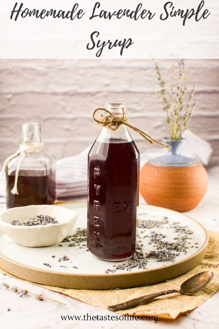 A glass bottle of dark purple homemade lavender syrup with a twine bow sits on a plate surrounded by dried lavender buds. Another bottle, a small bowl of lavender, and a vase with lavender stems are in the background.