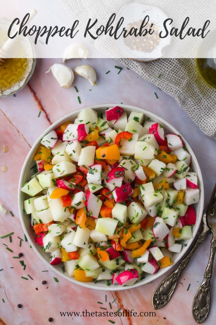 A bowl of chopped kohlrabi salad with colorful vegetables, garnished with herbs. Garlic, olive oil, and a cloth napkin are nearby. Text reads Chopped Kohlrabi Salad and www.thetastesoflife.com.