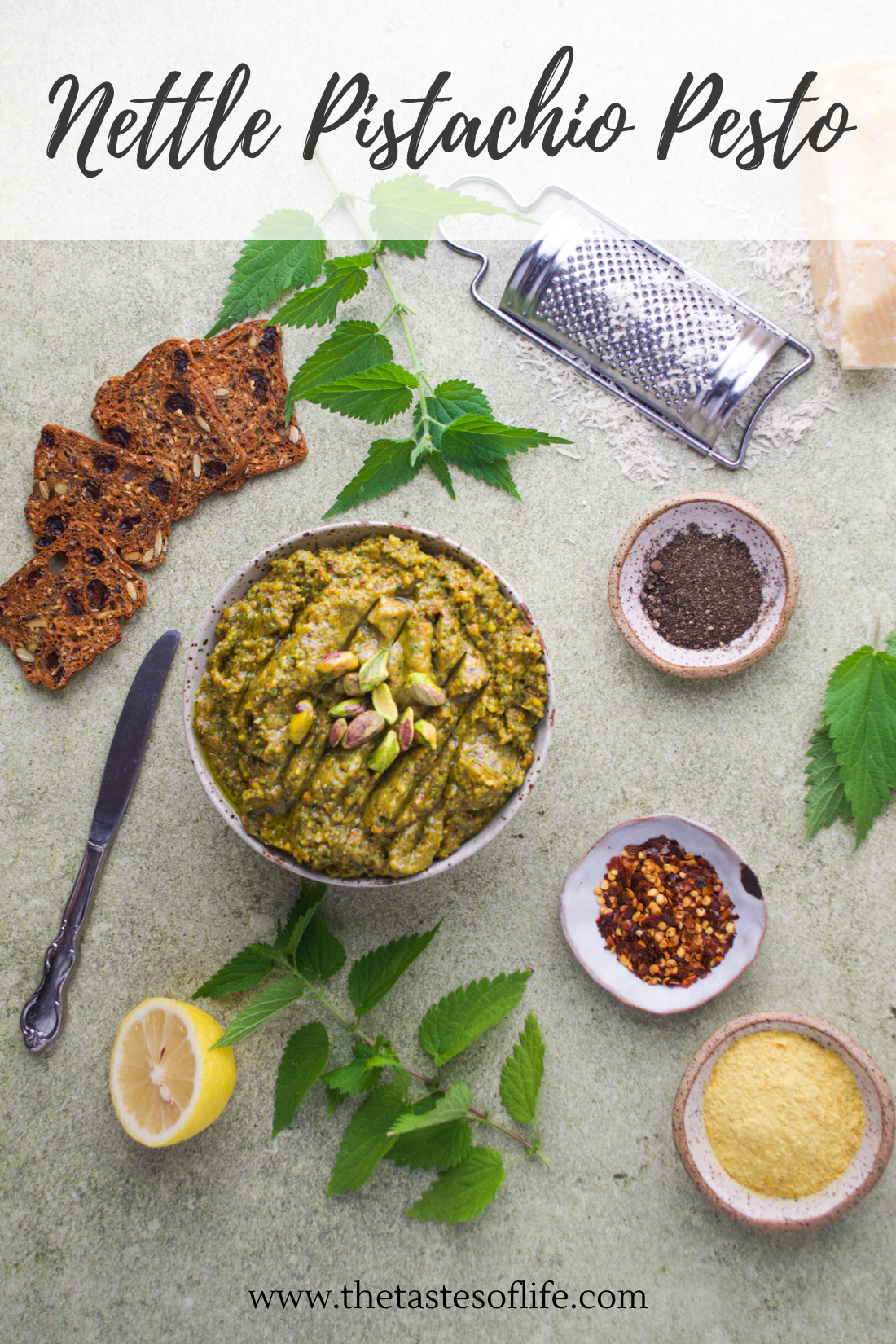 Bowl of nettle pistachio pesto surrounded by bread, nettle leaves, lemon, spices, grated cheese, and a grater on a light surface. Website text at the bottom reads “thetasteoflife.com.”.