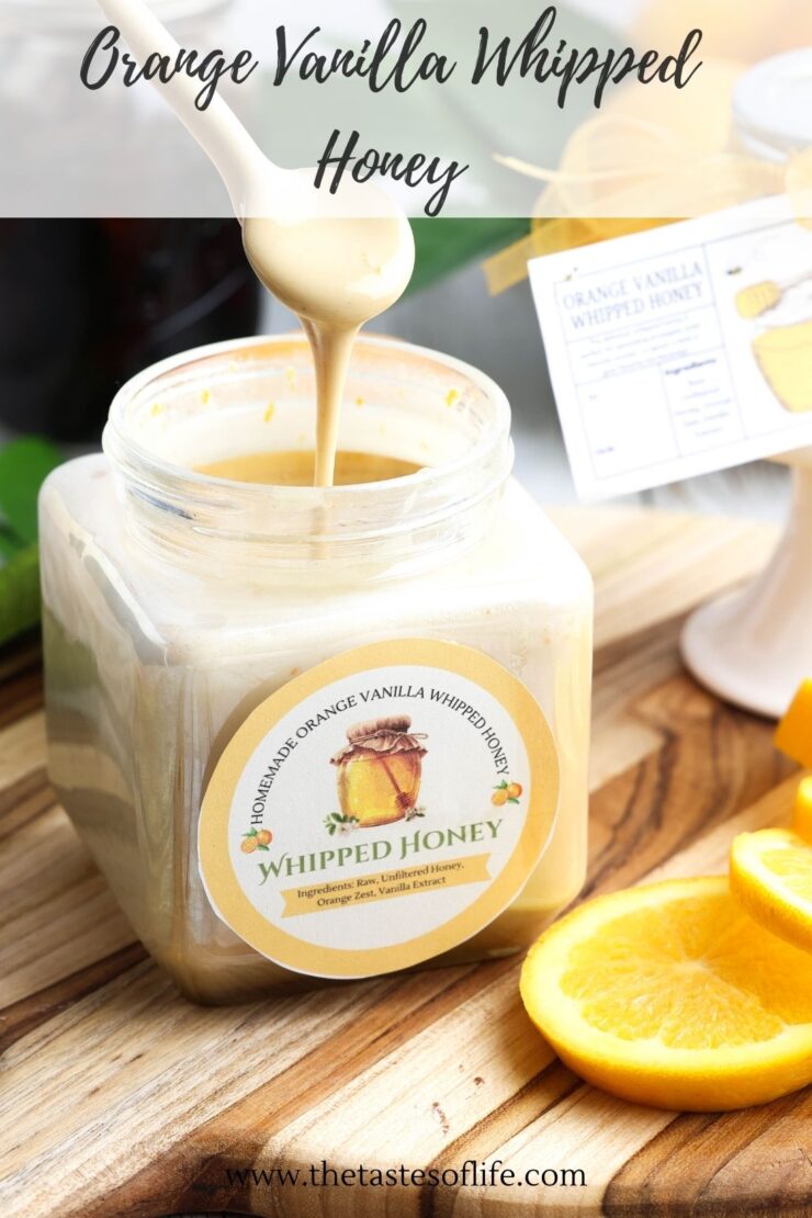 A spoonful of orange vanilla whipped honey being drizzled into a square glass jar with a label reading Homemade Orange Vanilla Whipped Honey, set on a wooden board next to orange slices.