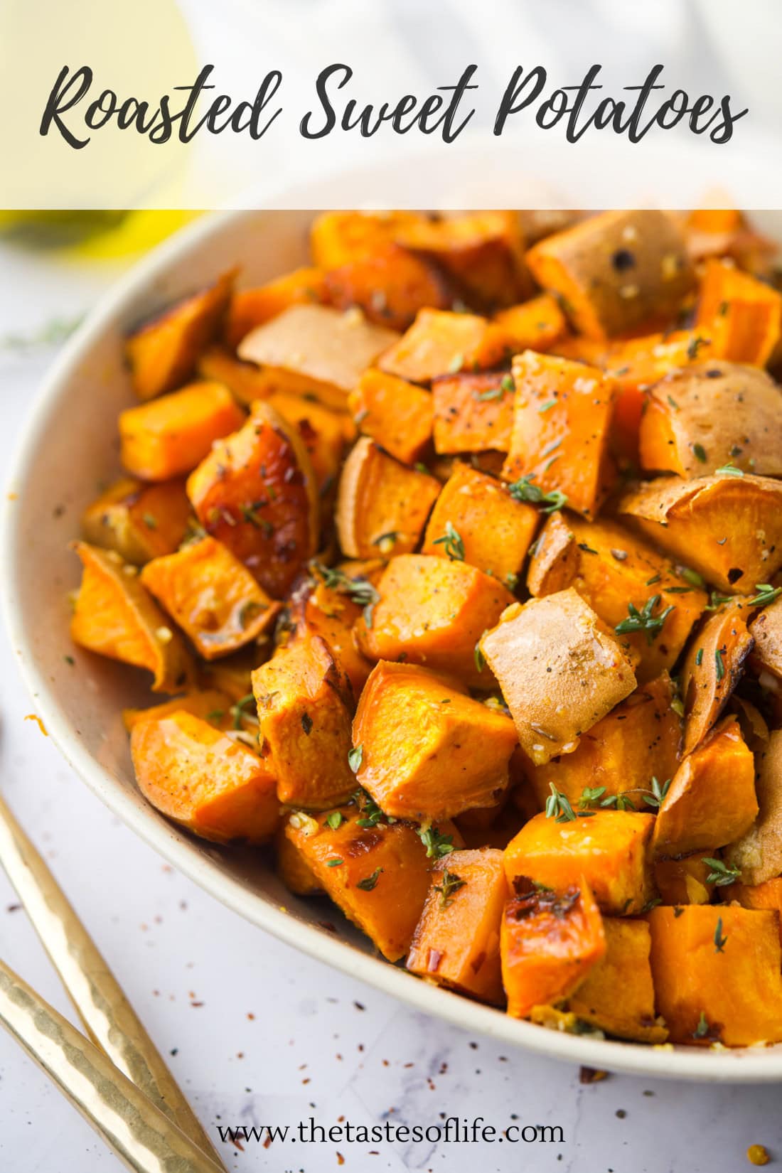 A bowl of roasted sweet potato cubes garnished with herbs, served on a white plate with two gold forks beside it.