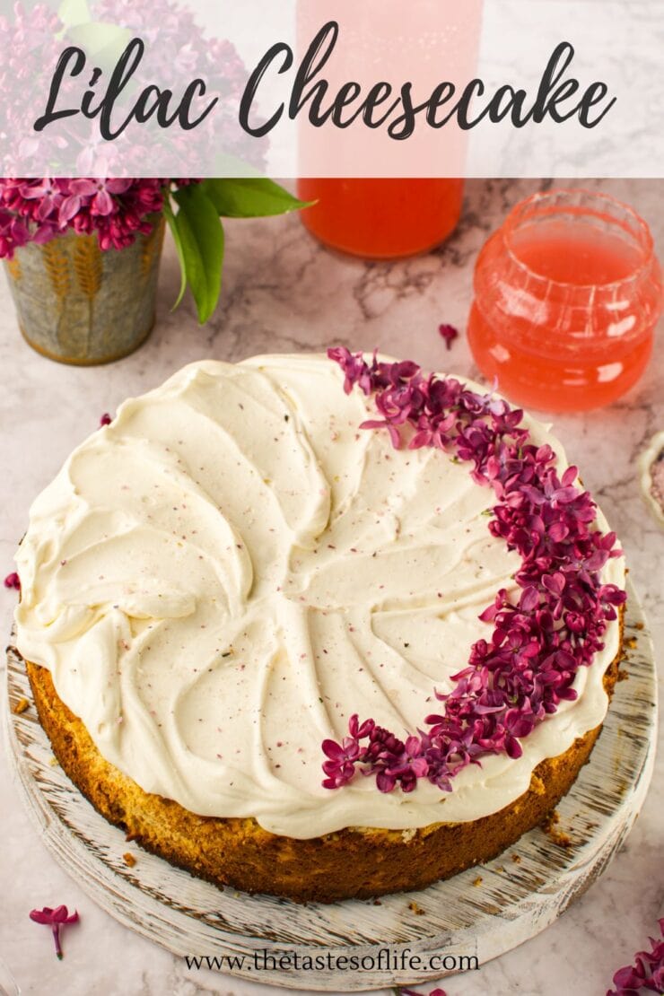 A cheesecake topped with creamy frosting and decorated with a cluster of purple lilac flowers, set on a light marble surface with lilac flowers and pink drinks in the background. Text reads &ldquo;Lilac Cheesecake.&rdquo;.