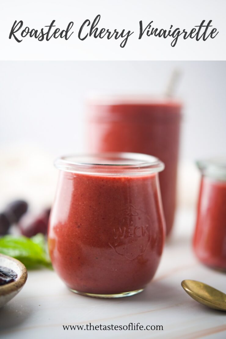 A glass jar filled with red roasted cherry vinaigrette sits in focus, with more jars of the same vinaigrette and a spoon blurred in the background. The words Roasted Cherry Vinaigrette are at the top.