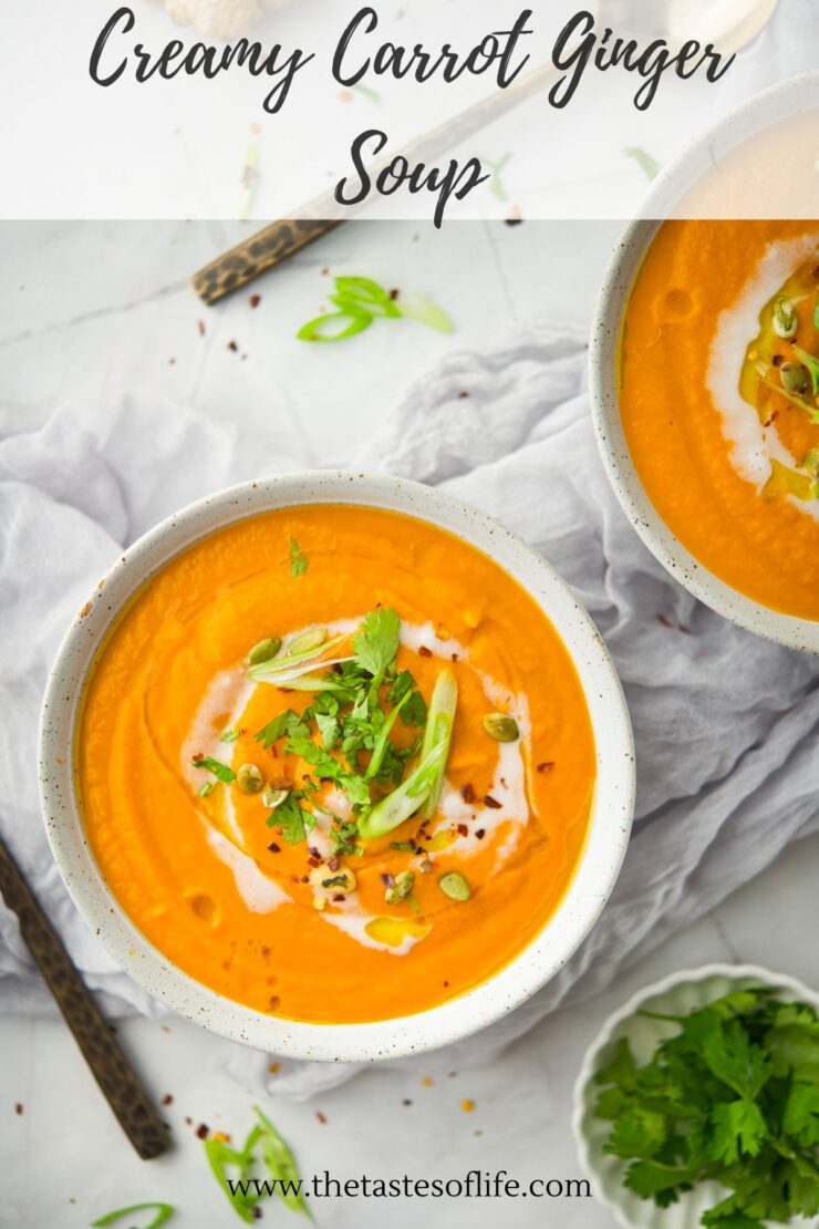 A bowl of creamy carrot ginger soup topped with cream, herbs, scallions, and seeds, set on a white surface with a spoon and garnishes nearby.