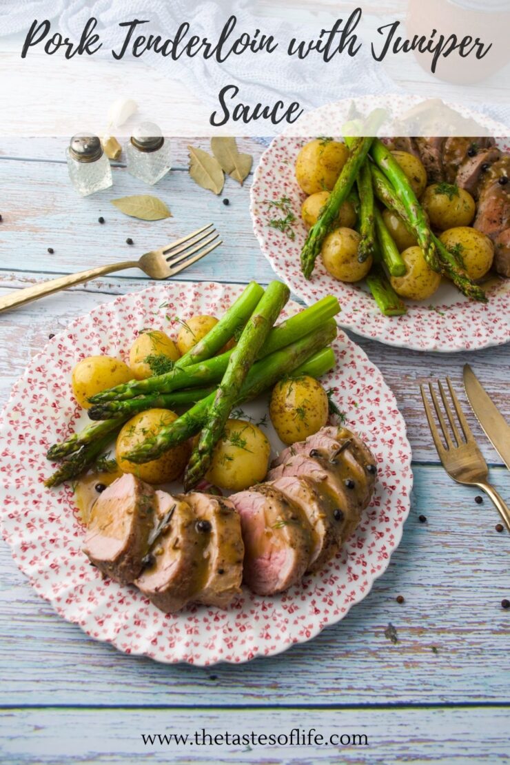 Sliced pork tenderloin with juniper sauce, baby potatoes, and asparagus on a floral plate, accompanied by a fork and knife on a rustic blue wooden table.