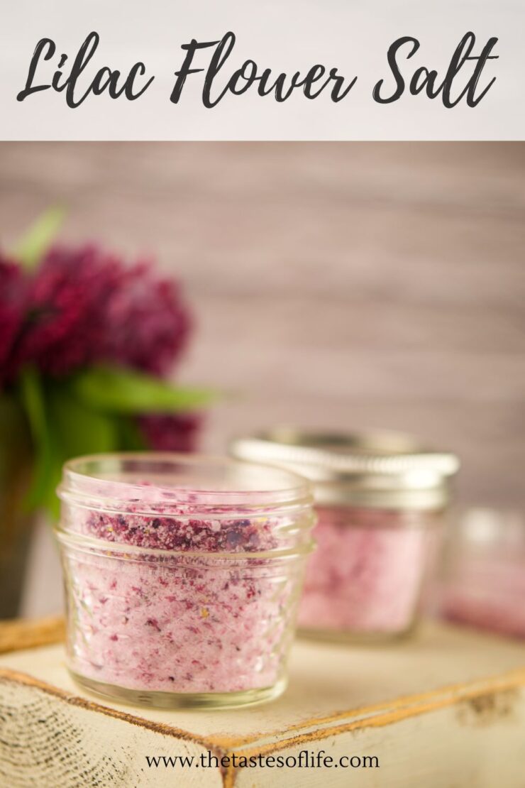 A small glass jar filled with lilac flower salt sits on a wooden surface, with another jar and a vase of lilac flowers blurred in the background. Text above reads Lilac Flower Salt.