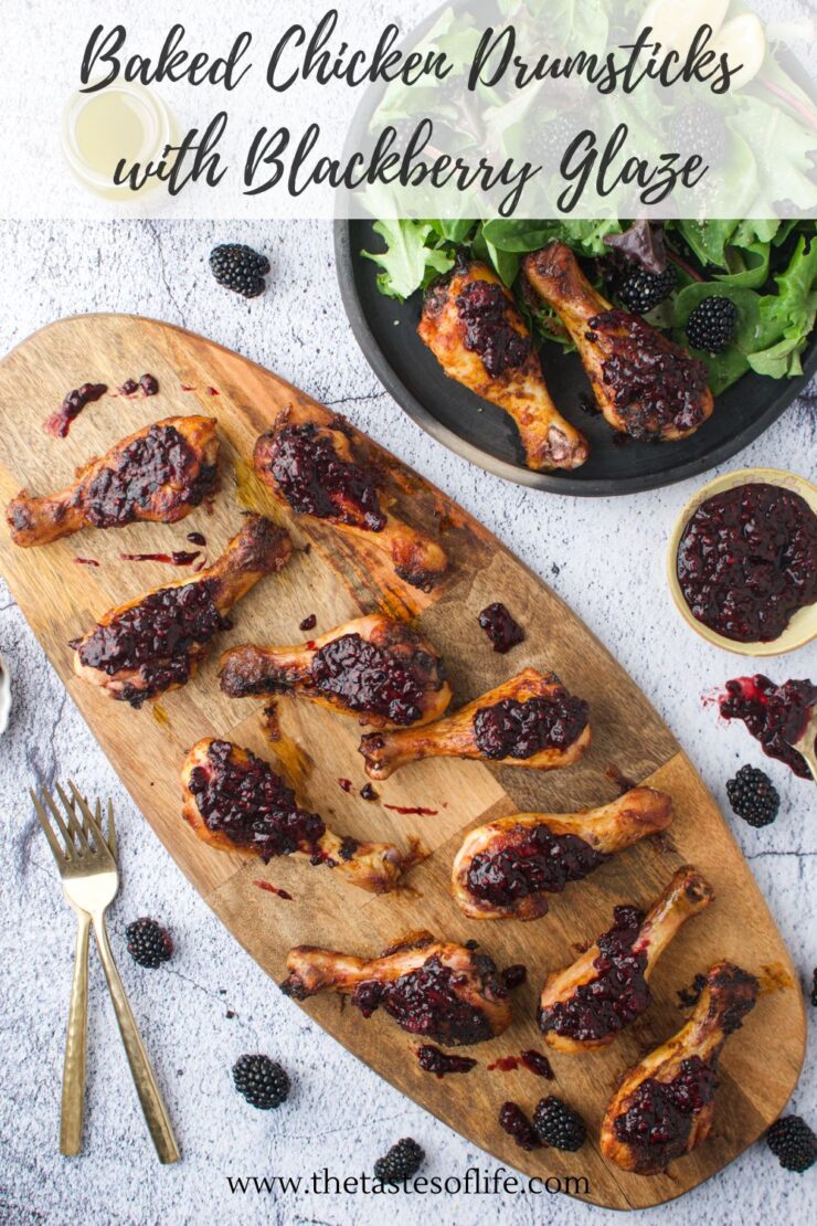 Overhead view of baked chicken drumsticks topped with blackberry glaze on a wooden board. A bowl of salad, fresh blackberries, forks, and a small bowl of glaze are nearby. Text at the top reads, Baked Chicken Drumsticks with Blackberry Glaze.
