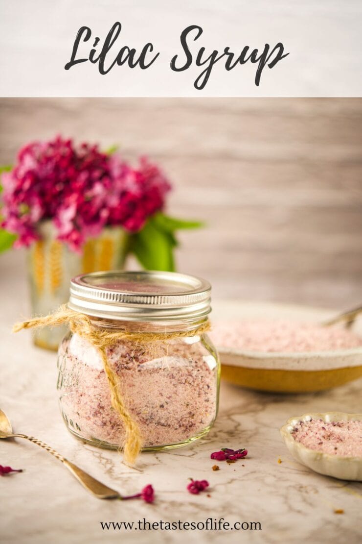 A glass jar filled with pink lilac syrup powder, tied with twine, sits on a light surface. Behind it are a bowl of the same powder, a spoon, and a bouquet of lilac flowers. Text reads Lilac Syrup at the top.
