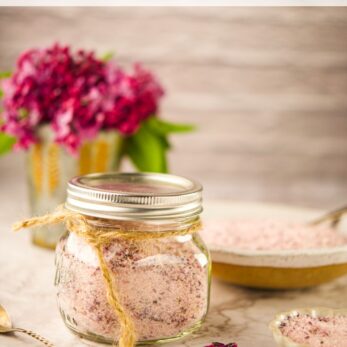 A glass jar filled with lilac syrup sits on a table, surrounded by a bowl and spoon containing more syrup. Purple lilac flowers are blurred in the background. The text Lilac Syrup appears at the top.