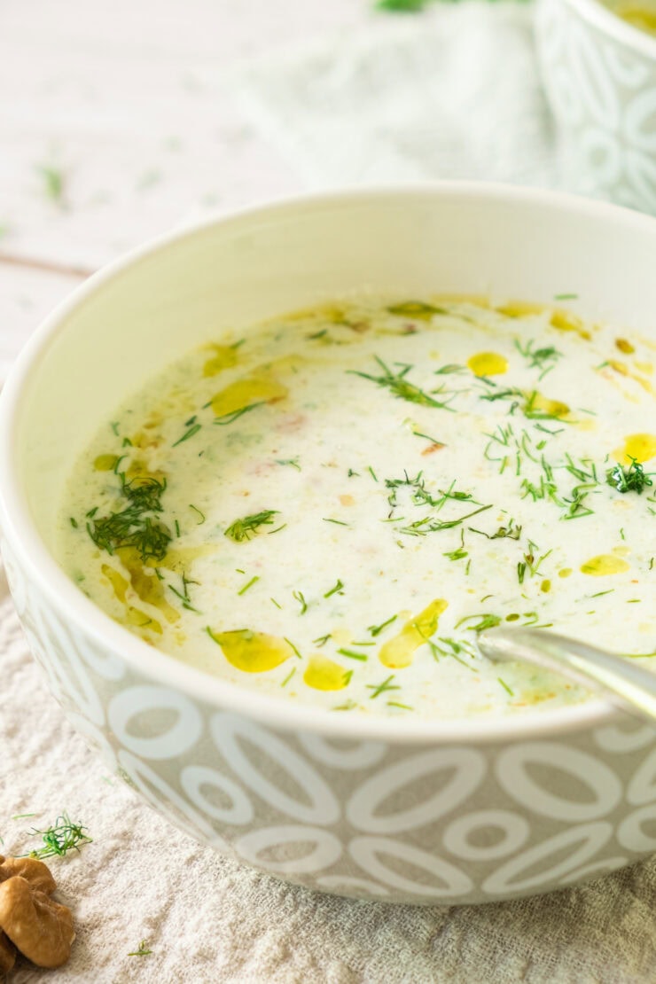 A bowl of creamy white soup garnished with fresh chopped dill and a drizzle of olive oil, placed on a textured napkin with a spoon inside the bowl.