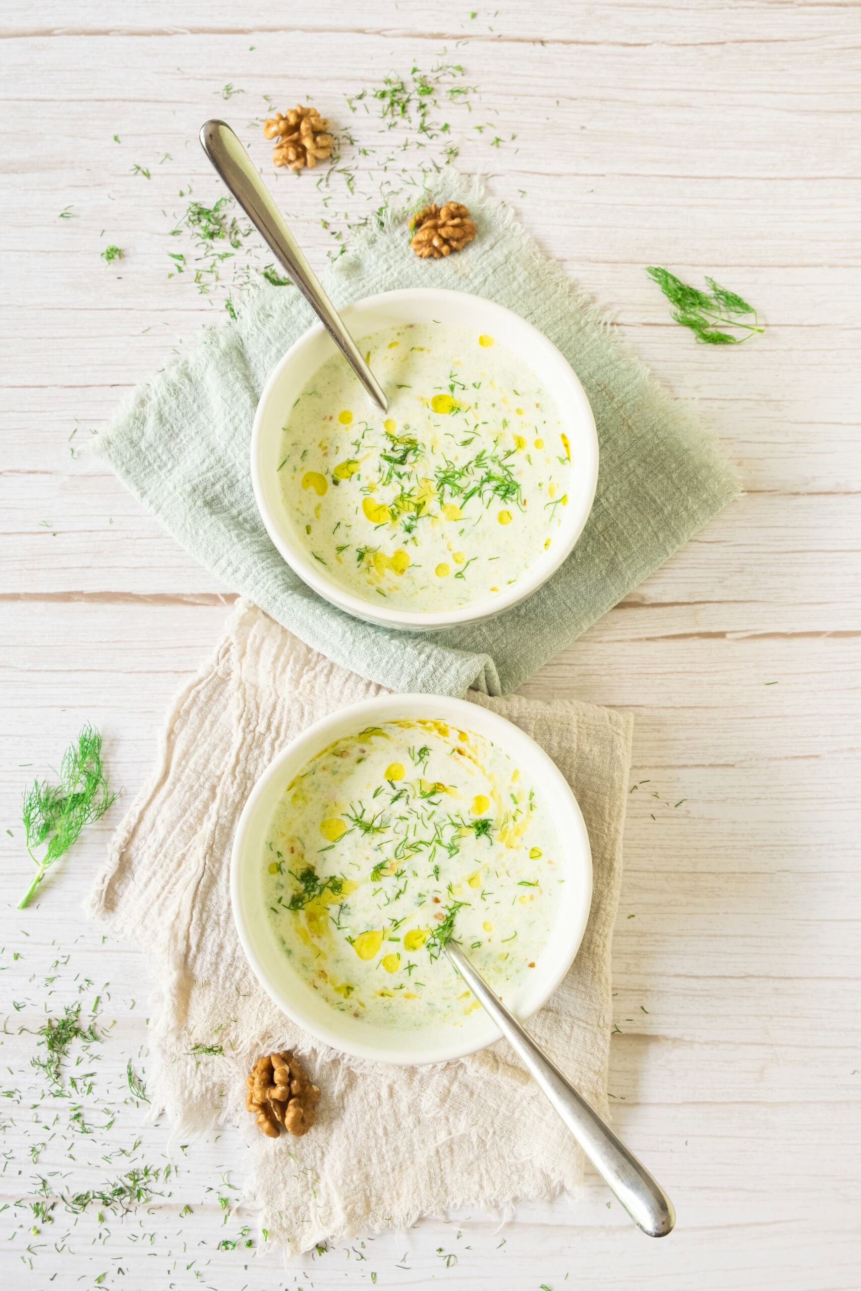 Two bowls of creamy soup garnished with herbs and a drizzle of oil, each with a spoon, placed on light cloth napkins on a white wooden surface. Pieces of fresh dill and walnuts are scattered around the bowls.