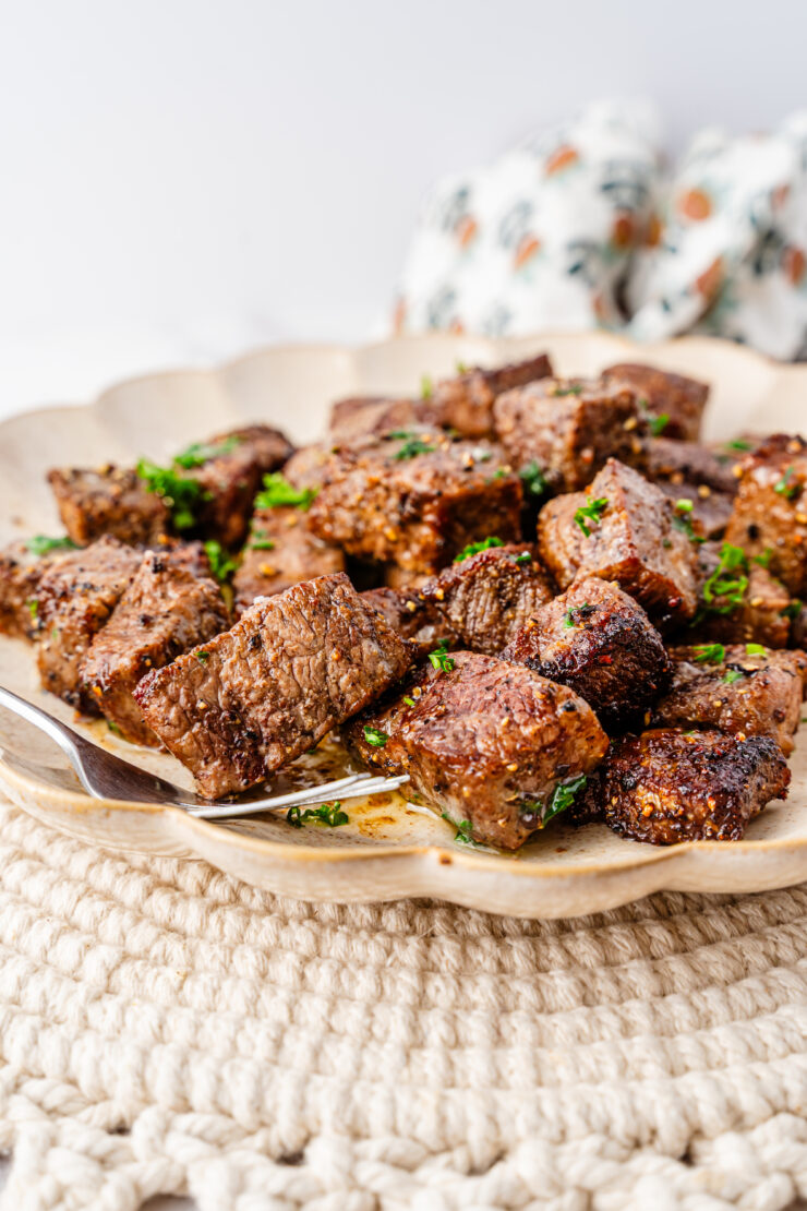 A plate filled with browned, Air Fryer Steak Bites garnished with chopped parsley, resting on a textured tan placemat with a fork on the side.