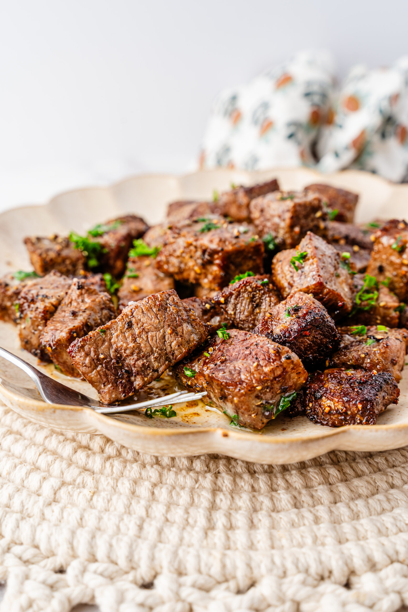 A plate filled with browned, Air Fryer Steak Bites garnished with chopped parsley, resting on a textured tan placemat with a fork on the side.