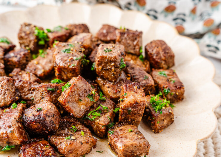A plate of seared steak bites garnished with chopped parsley, served on a beige dish with a patterned cloth in the background.