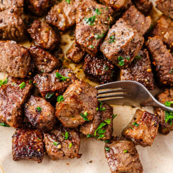 A plate of Air Fryer Steak Bites garnished with chopped parsley, with a fork picking up a few pieces. The steak looks juicy and well-seasoned, served on a cream-colored textured plate.