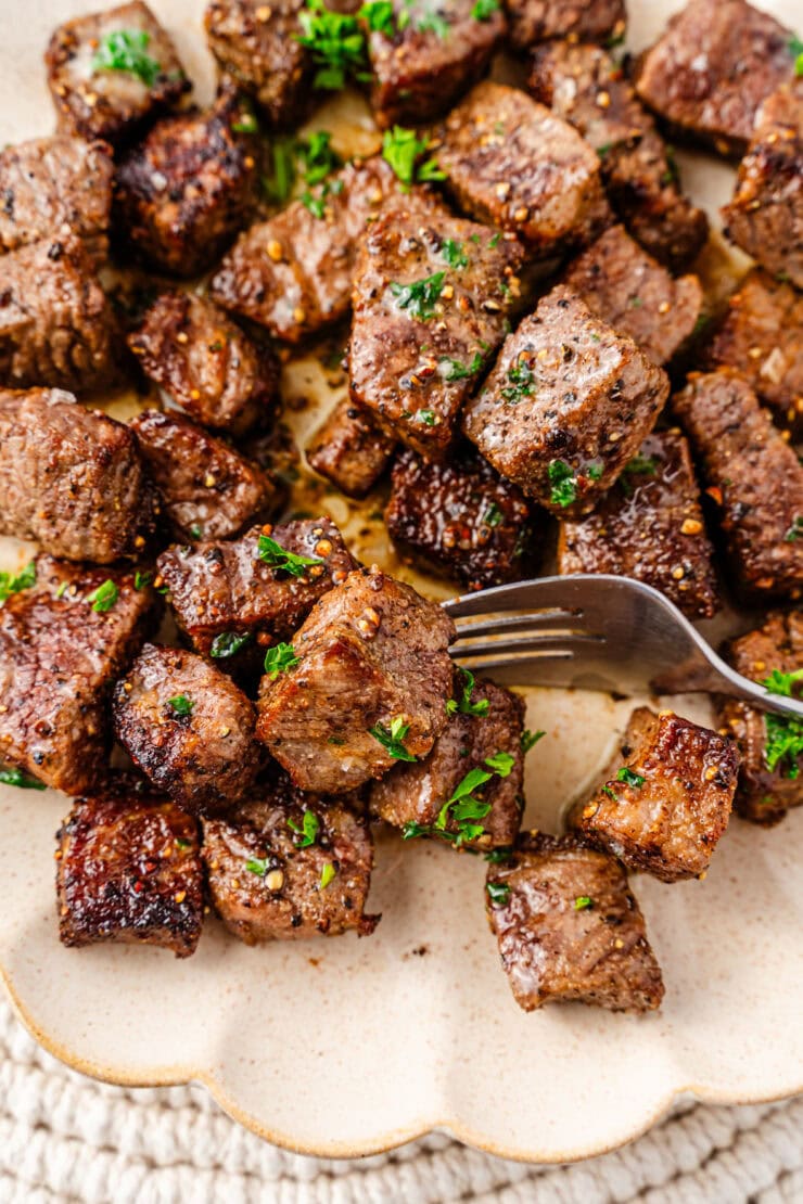 A plate of Air Fryer Steak Bites garnished with chopped parsley, with a fork picking up a few pieces. The steak looks juicy and well-seasoned, served on a cream-colored textured plate.