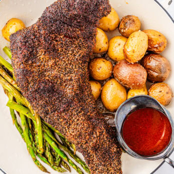 A Brisket style smoked tri tip with roasted baby potatoes, grilled asparagus, and a small bowl of red sauce served in a white dish.
