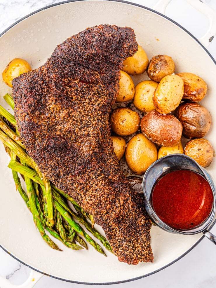 A Brisket style smoked tri tip with roasted baby potatoes, grilled asparagus, and a small bowl of red sauce served in a white dish.