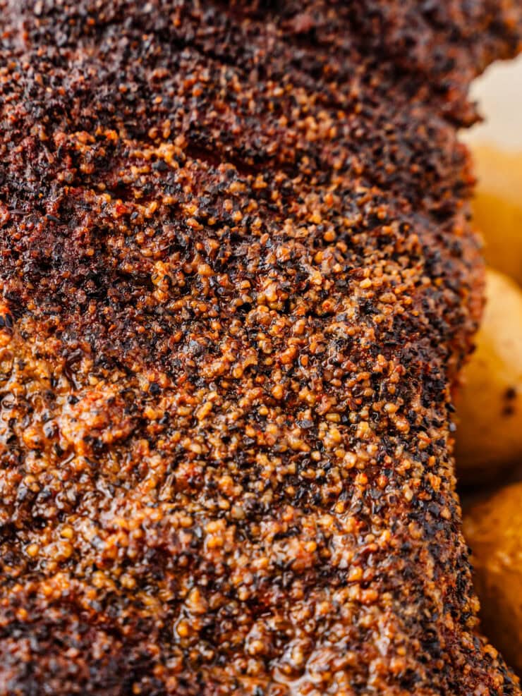 Close-up of a heavily seasoned and crusted piece of cooked meat, showing a textured spice rub surface with visible granules and a glistening finish. Some yellow potatoes are partially visible in the background.
