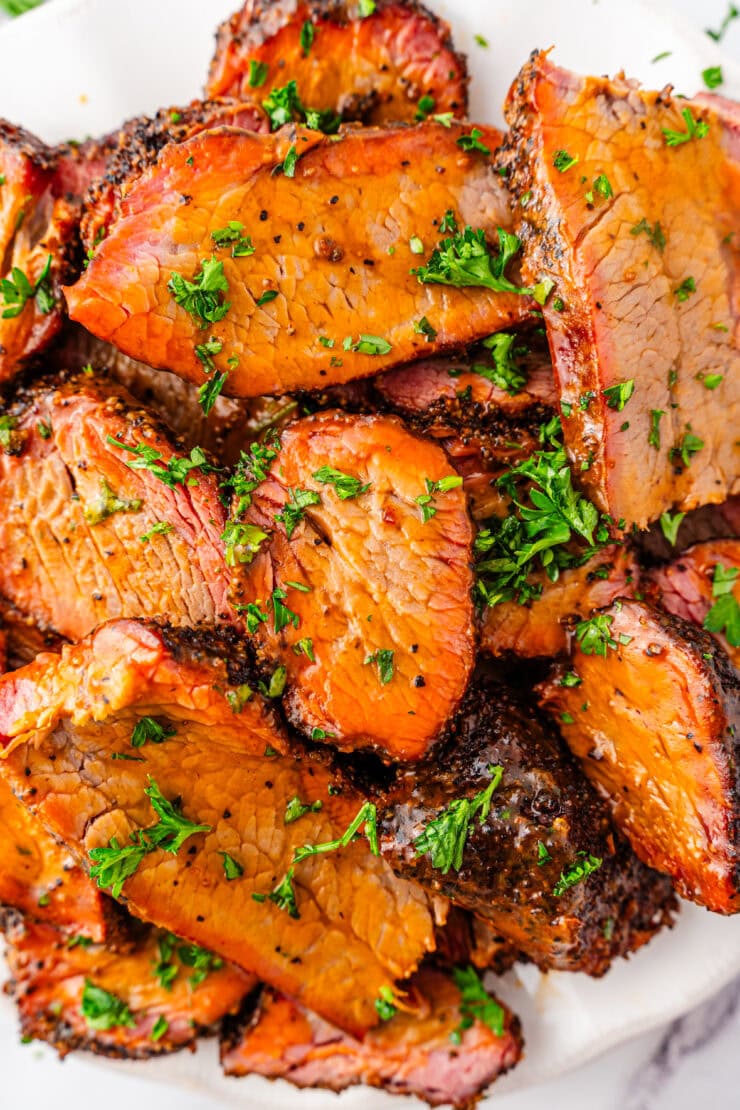Slices of smoked brisket with a dark, seasoned crust are piled on a plate and garnished with chopped fresh parsley. The meat is juicy with a visible smoke ring.