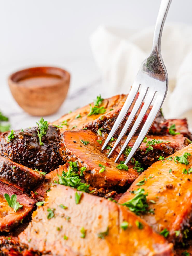 A fork pierces a slice of juicy, seasoned roast beef garnished with chopped parsley, with a small bowl of sauce in the background.