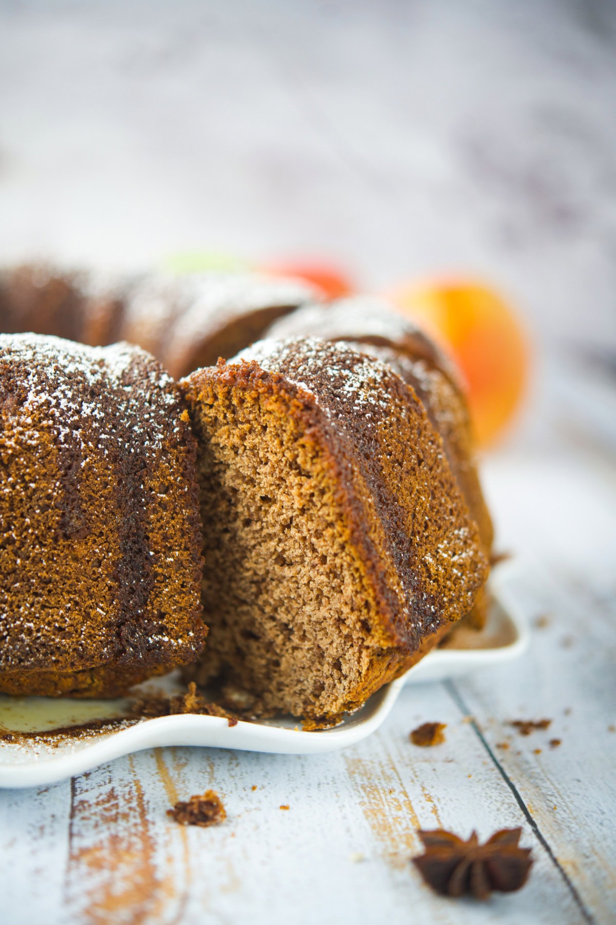 A sliced bundt cake dusted with powdered sugar sits on a white plate, revealing its soft, brown interior. The background is blurred, showing subtle hints of fruit and spices.