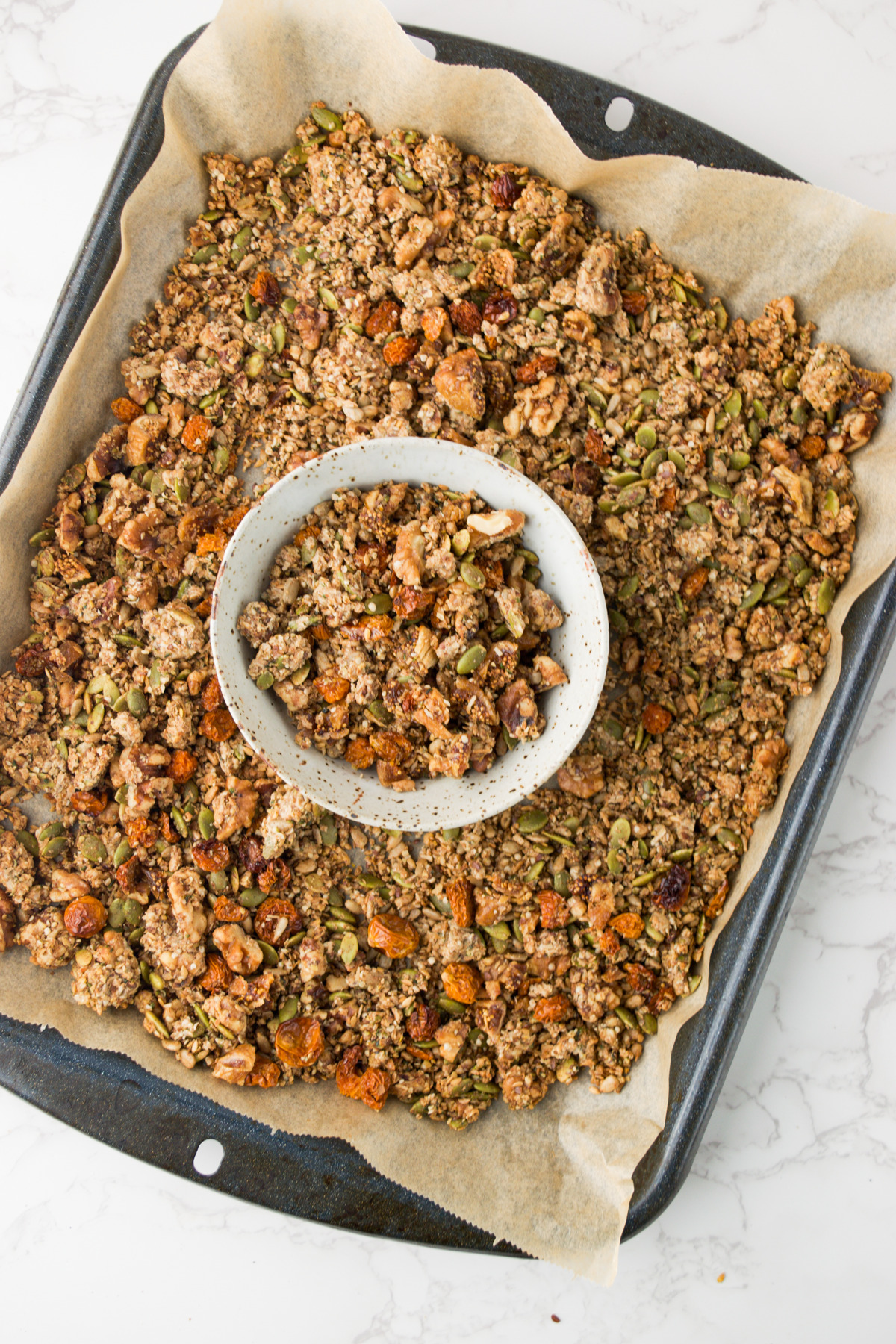 A baking tray lined with parchment paper holds a batch of homemade granola. A small bowl with granola sits on top of the tray. The granola contains oats, seeds, and nuts, and is spread in clusters.