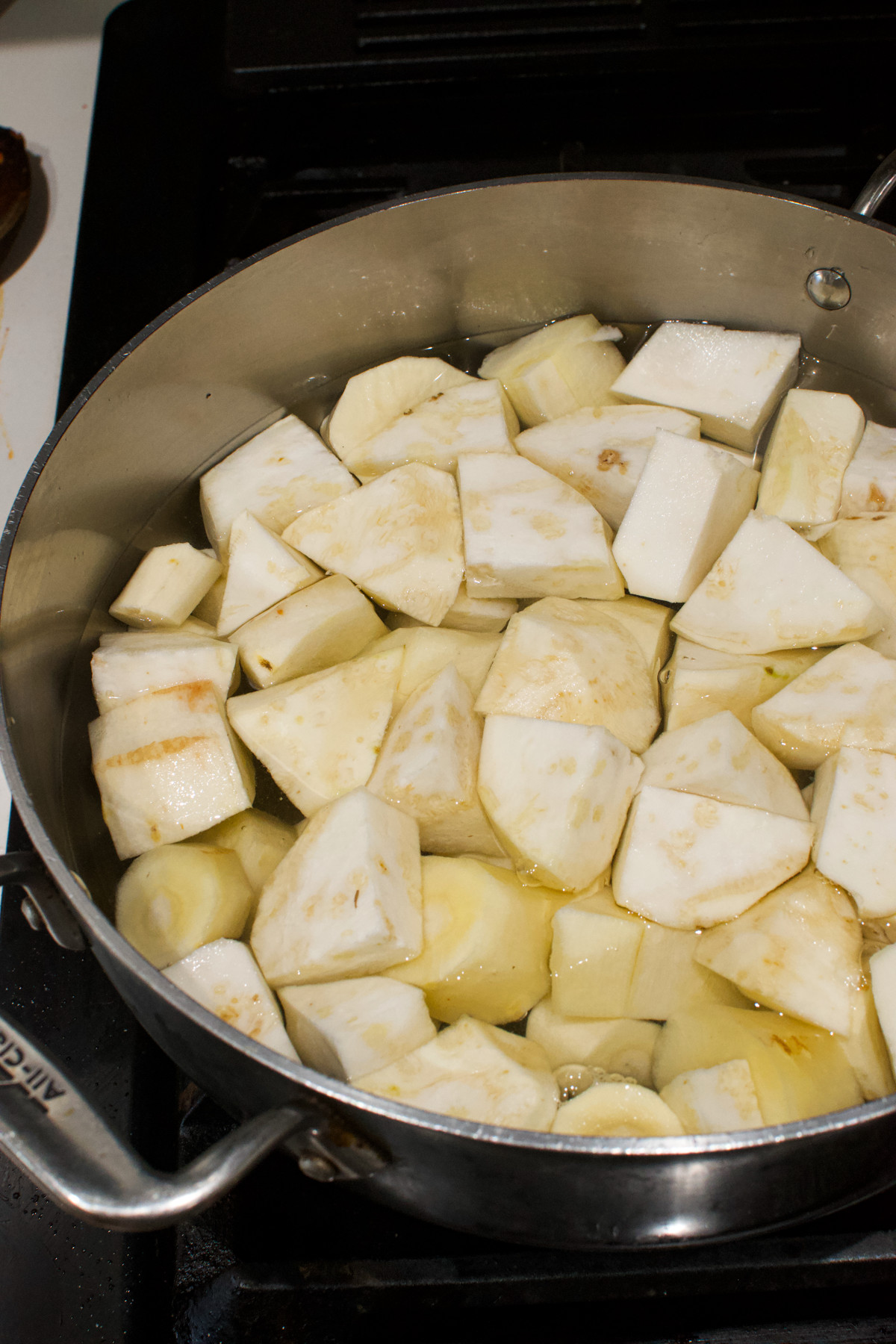 Large chunks of peeled celery root are simmering in oil or water in a stainless steel pan on a stovetop.