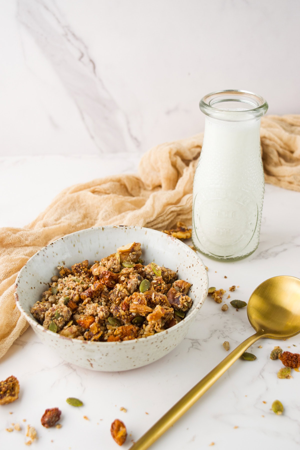 A bowl of granola with nuts and seeds sits on a white marble surface next to a vintage-style glass bottle of milk, a gold spoon, and a beige cloth.