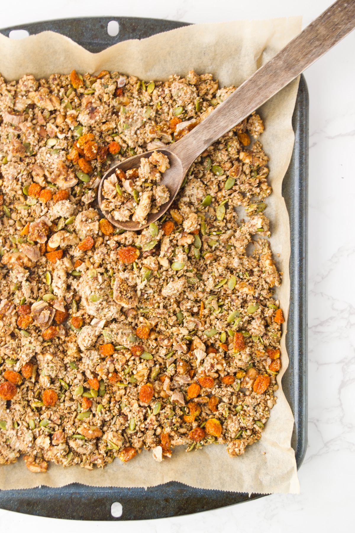 A baking tray lined with parchment paper holds a freshly baked granola mix with oats, seeds, nuts, and orange dried fruit pieces. A wooden spoon rests on the tray, scooping some granola.