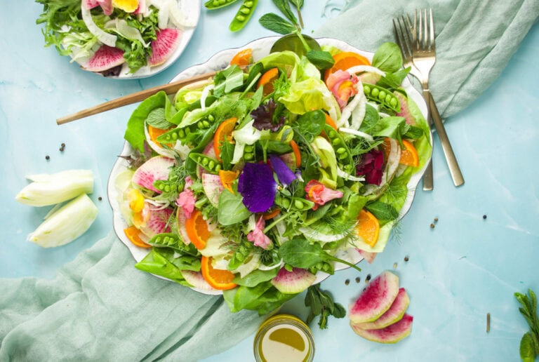 Vibrant spring salad on a white plate featuring a colorful mix of fresh greens, snap peas, shaved fennel, carrots, edible flowers, and herbs. The salad is set on a soft blue background with a napkin underneath and a fork in the foreground, evoking a fresh, seasonal, and garden-to-table feel