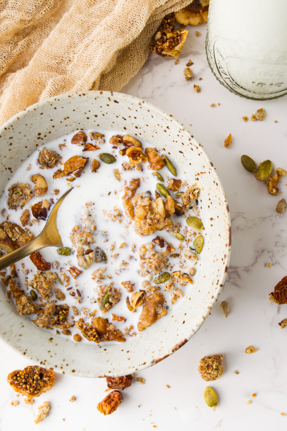 A speckled ceramic bowl filled with milk and granola sits on a marble surface, with a spoon inside. Pieces of granola and seeds are scattered around, and a glass of milk is nearby. A textured beige cloth is in the background.
