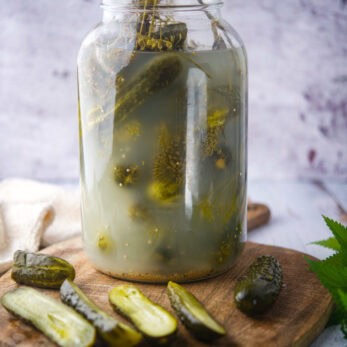 A large glass jar of pickles in brine sits on a wooden board with several sliced pickles and a knife beside it. The background is rustic and lightly textured.