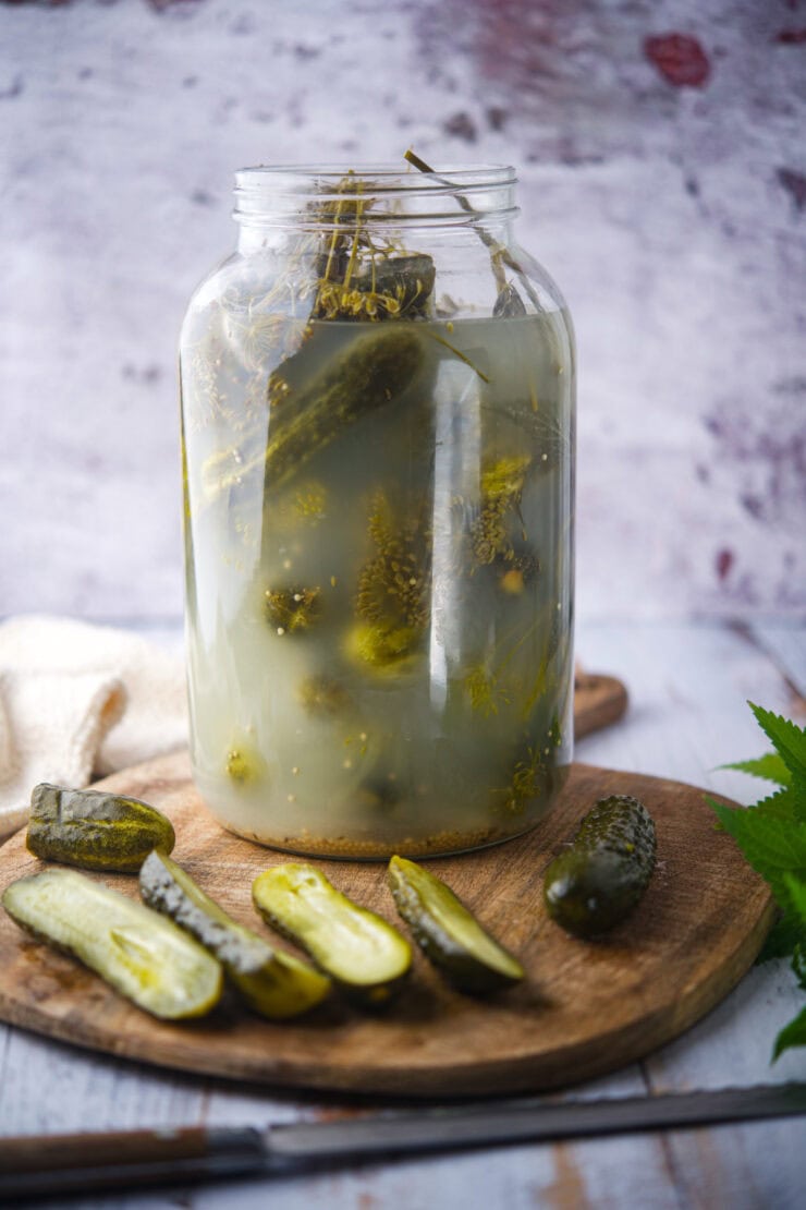 A large glass jar of pickles in brine sits on a wooden board with several sliced pickles and a knife beside it. The background is rustic and lightly textured.