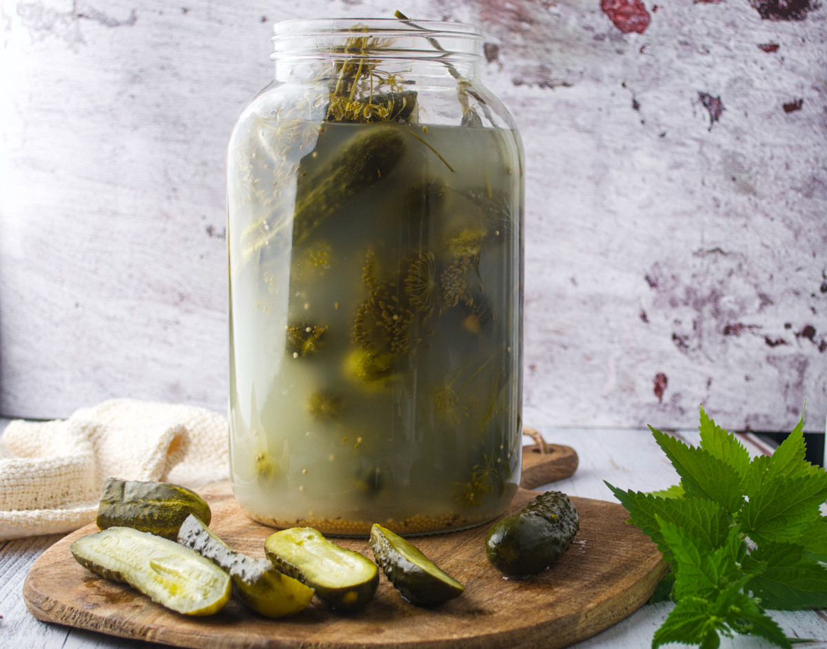 A large glass jar of pickles in cloudy brine sits on a wooden board with sliced pickles and a green leaf nearby. The background is a textured, light-colored wall.