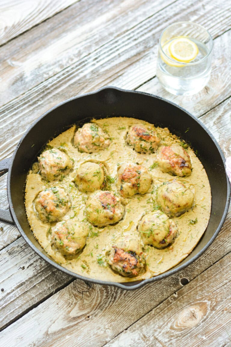 A black cast iron skillet filled with One-Pan Turkey Meatballs With Cashew Creamy Sauce, garnished with fresh herbs, sits on a rustic wooden table next to a silver serving spoon.