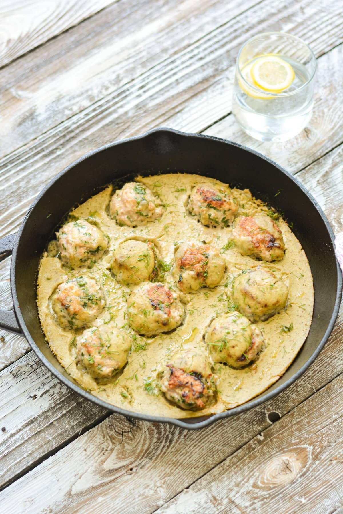 A black cast iron skillet filled with One-Pan Turkey Meatballs With Cashew Creamy Sauce, garnished with fresh herbs, sits on a rustic wooden table next to a silver serving spoon.
