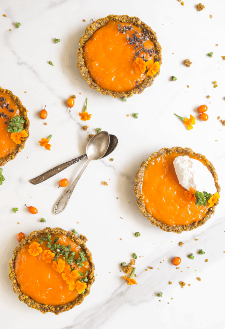 Top-down view of multiple seabuckthorn tartlets on a white marble background, with one partially eaten, garnished with edible flowers and accompanied by vintage spoons and scattered berries and herbs
