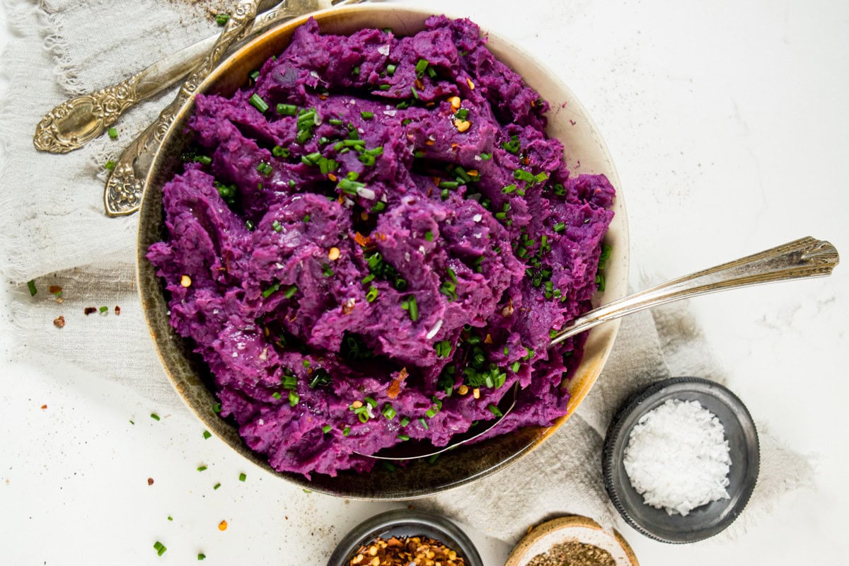 A bowl of vibrant purple mashed potatoes garnished with chopped chives, surrounded by small dishes of salt, pepper, and chili flakes, with a silver spoon and vintage cutlery on a white linen cloth.