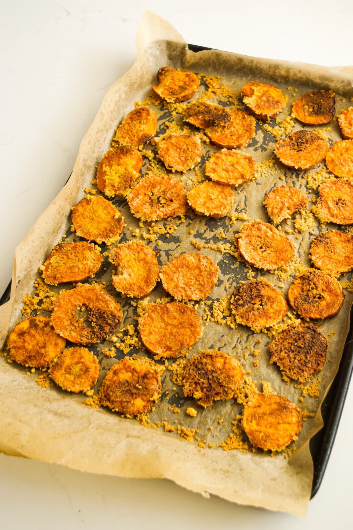 A baking tray lined with parchment paper holds several golden-brown, crispy, round chips spread out in a single layer, likely baked vegetable chips, on a white surface.