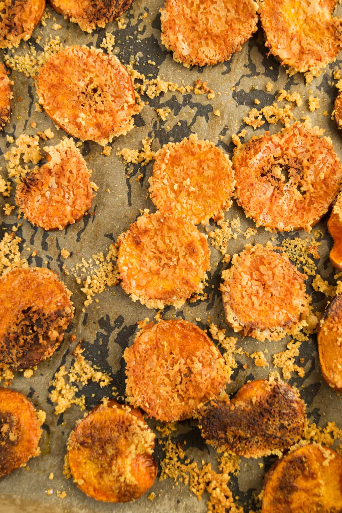 Baked, crispy carrot slices coated in breadcrumbs and cheese are spread out on a parchment-lined baking sheet. Some pieces are slightly browned, showing a crunchy texture.