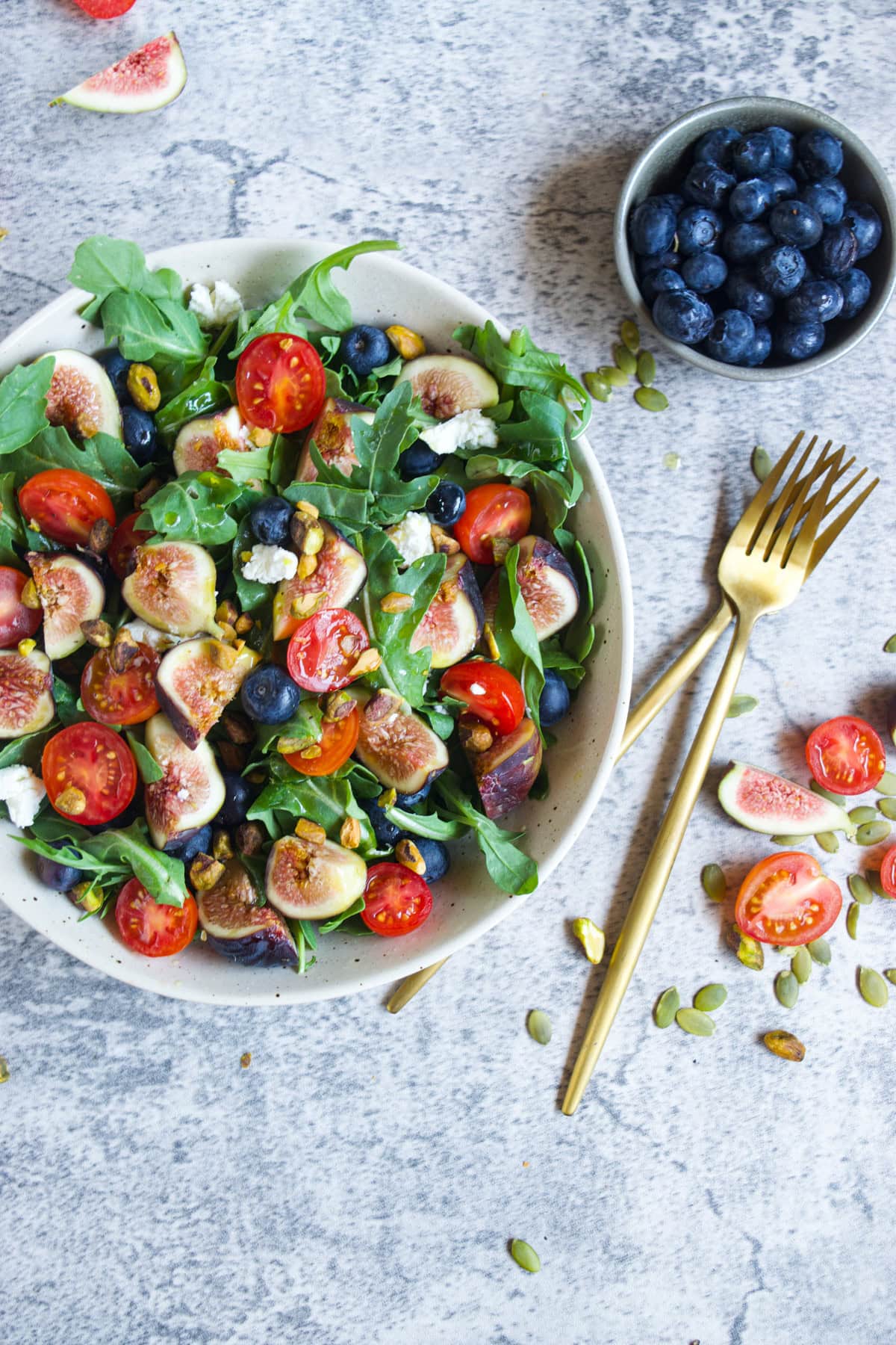 A fresh salad with arugula, cherry tomatoes, figs, blueberries, goat cheese, and nuts in a bowl beside a small dish of blueberries, with gold utensils on a gray textured surface.
