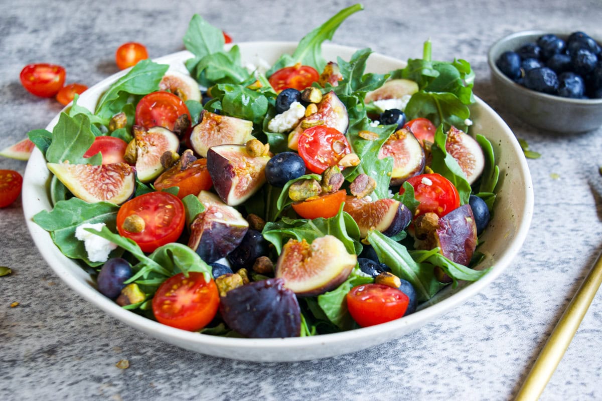 A bowl of fresh salad with arugula, halved cherry tomatoes, sliced figs, blueberries, and pistachios. A small bowl of blueberries sits in the background. The salad is arranged on a gray surface.