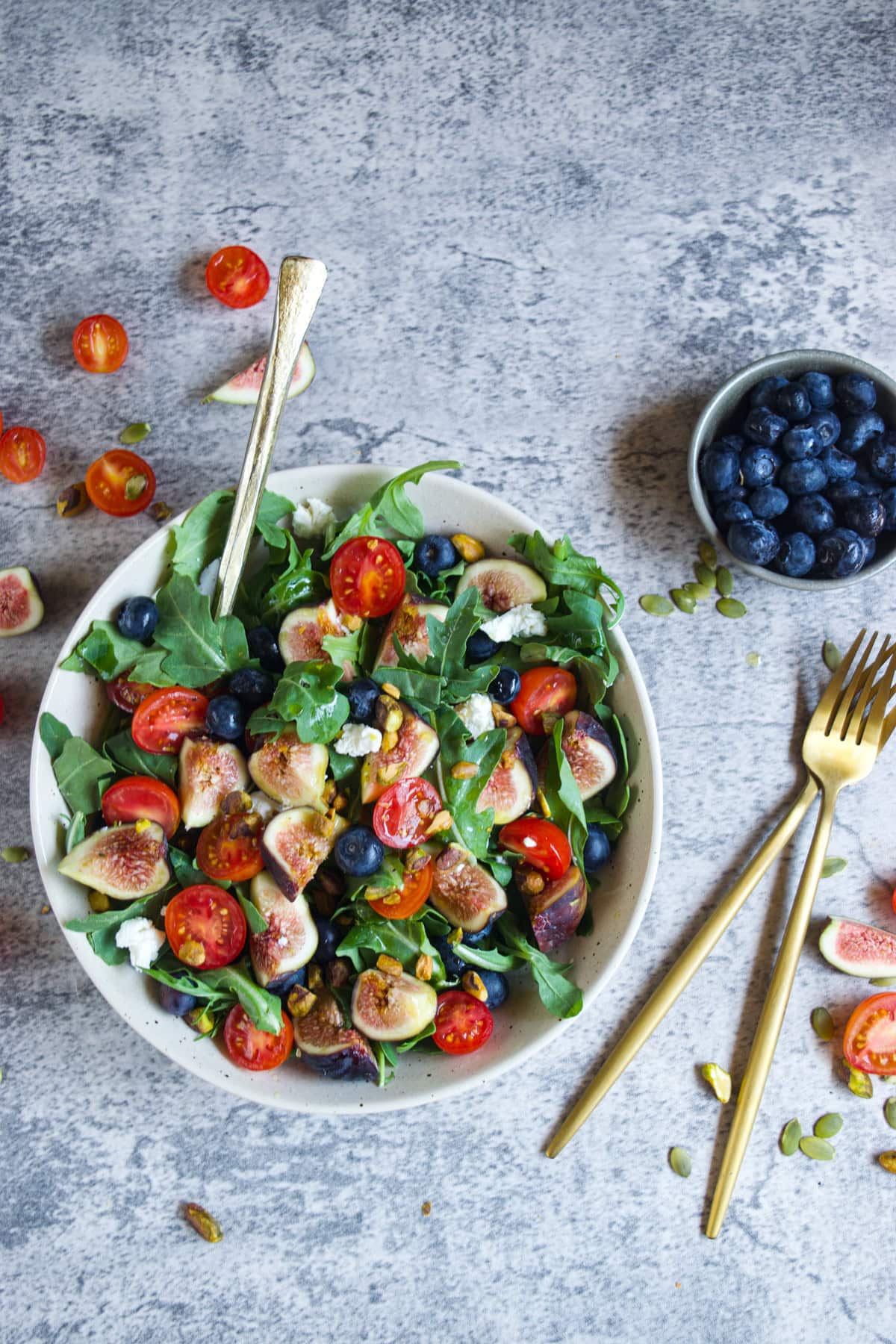 A bowl of salad with arugula, cherry tomatoes, figs, blueberries, and crumbled cheese. A gold fork is in the bowl, with another fork and knife nearby. A small bowl of blueberries and scattered ingredients surround the salad.