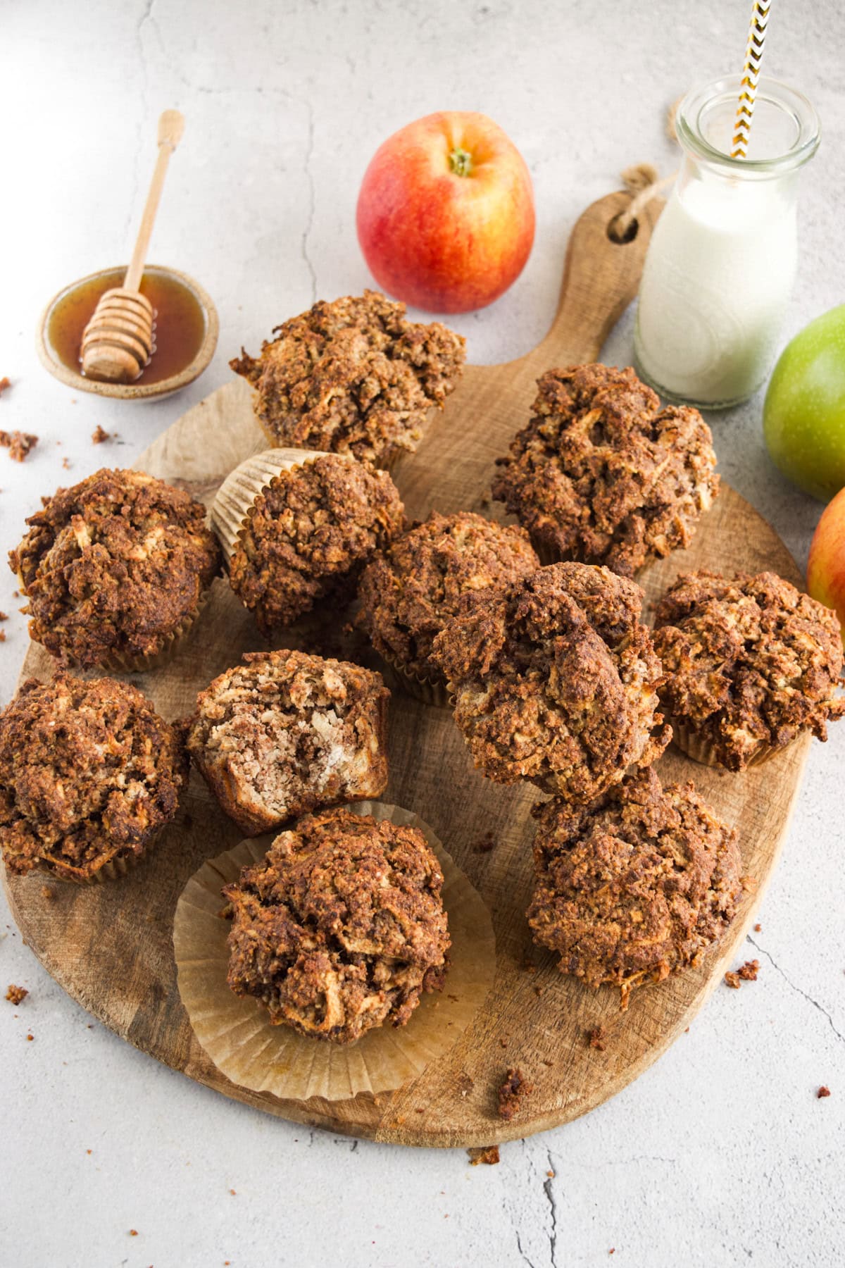 A wooden board with several bran muffins, one partially unwrapped and bitten, surrounded by apples, a jar of milk with a straw, and a dish of honey with a dipper on a light surface.