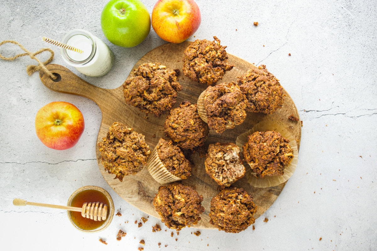 Overhead view of apple muffins on a round wooden board, surrounded by green and red apples, a jar of honey with a dipper, and a bottle of milk with a string bow on a light textured surface.