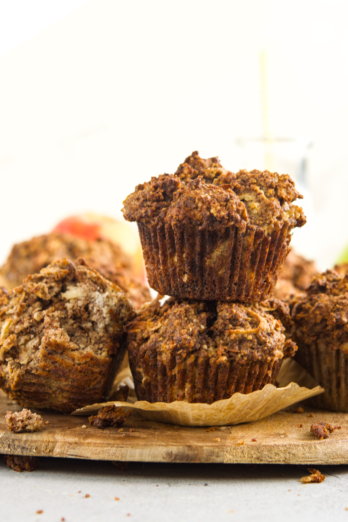 Two cinnamon muffins stacked on top of each other on a wooden board, with more muffins and muffin crumbs scattered around. The background is softly blurred.