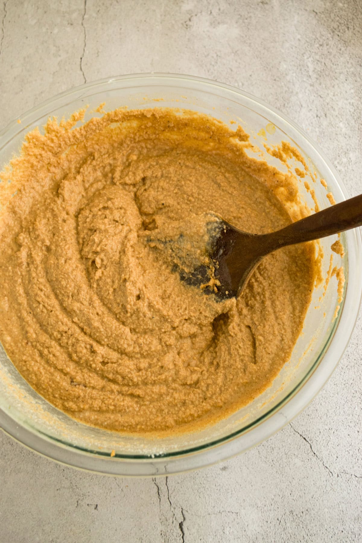 A glass bowl filled with light brown batter is being mixed with a wooden spoon on a light gray textured surface.