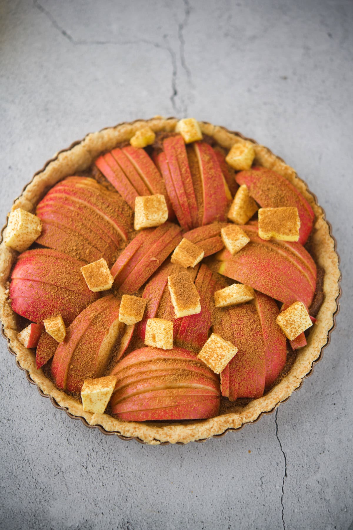 A close-up of an apple tart topped with thinly sliced red apples, sprinkled with cinnamon, and decorated with small cubes of toasted bread or pastry, set on a textured gray surface.